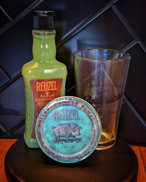 Reuzel pomade and scrub shampoo beside an American Barber Company glass